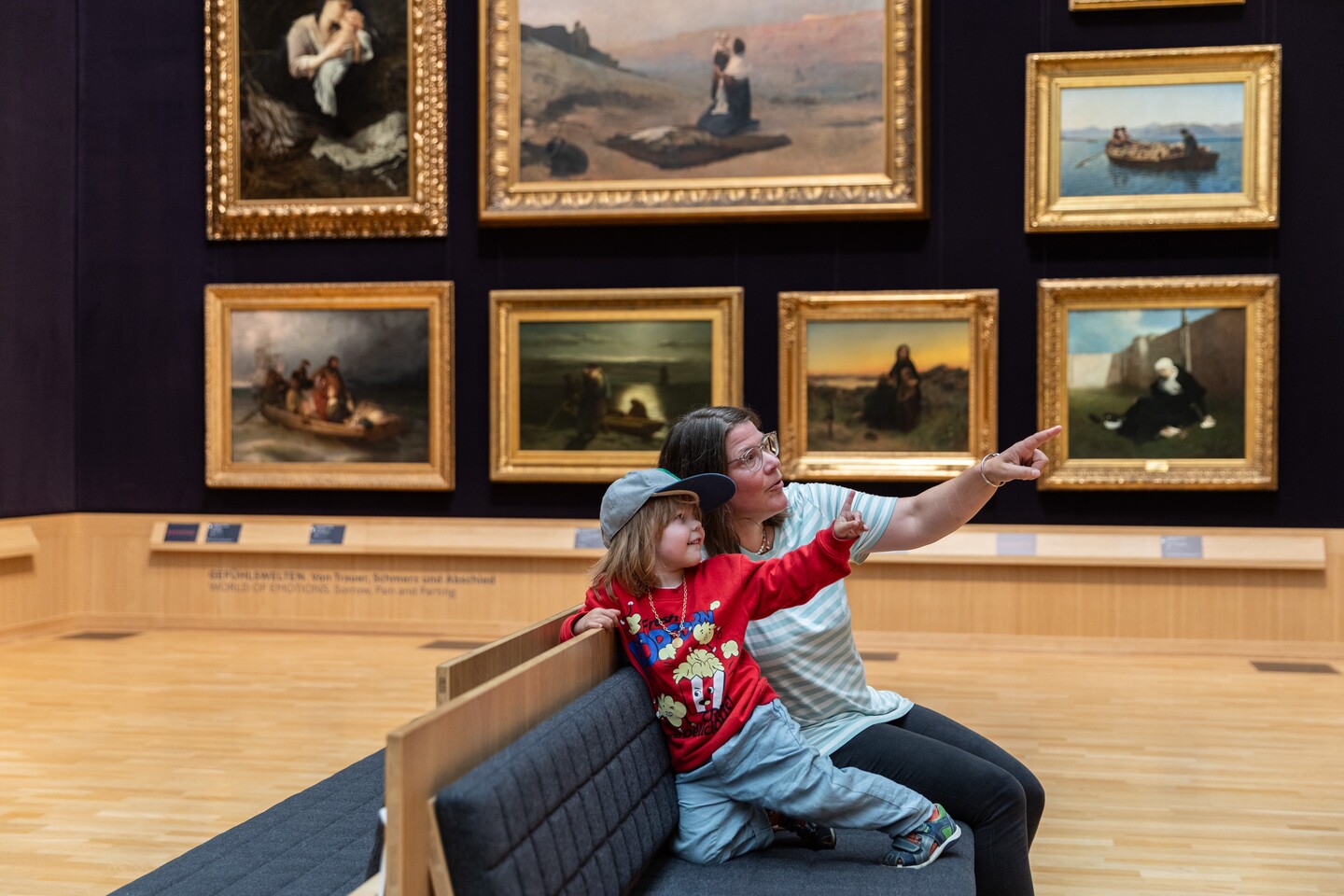 Mutter und Sohn sitzen auf einer Bank im Ausstellungsraum und zeigen auf Gemälde